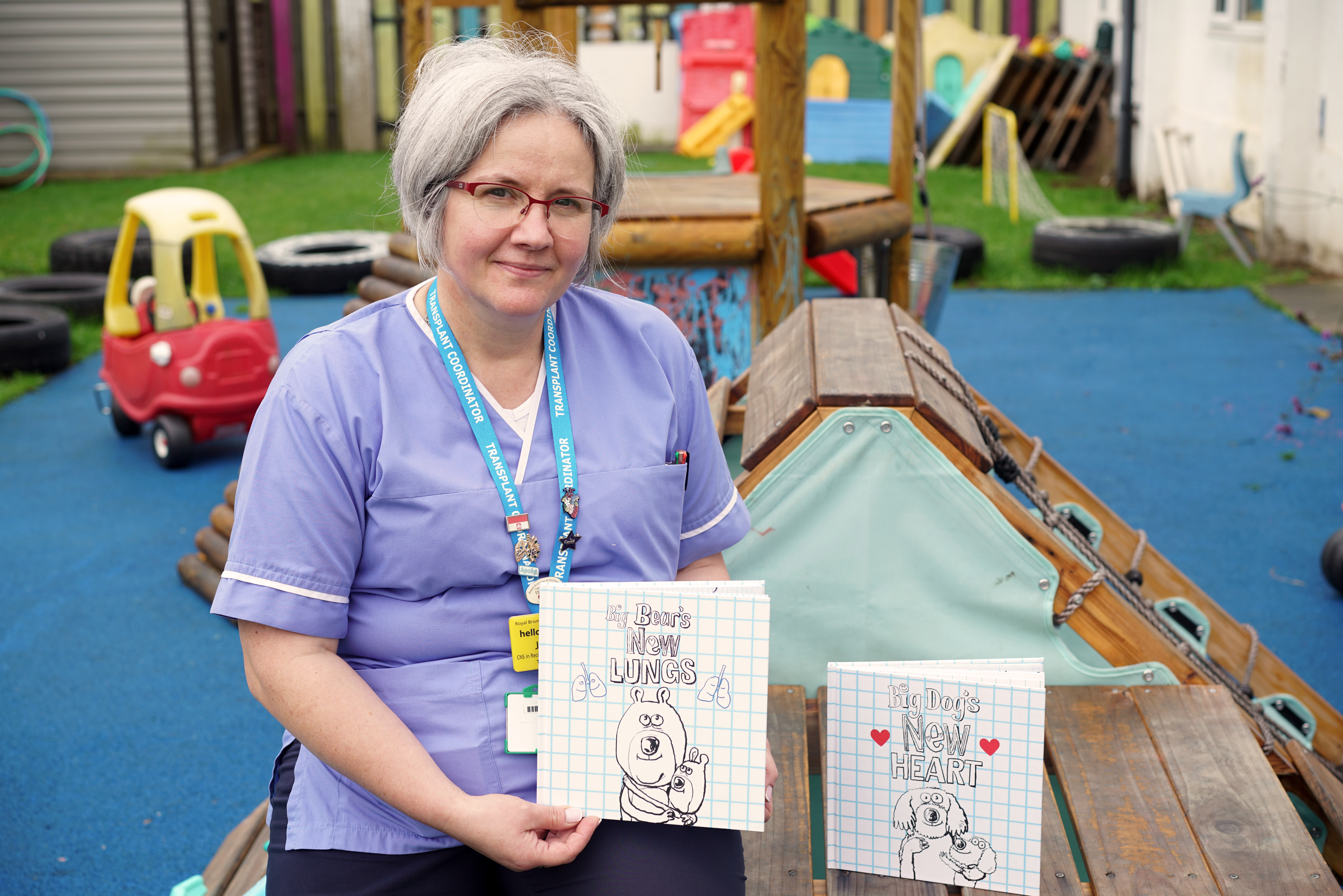 Image of Jola holding a transplant activity book. She is sitting in an outdoor area of a nursery. Jola is wearing a nurse's uniform, glasses and a blue lanyard. Next to her is another transplant activity book.